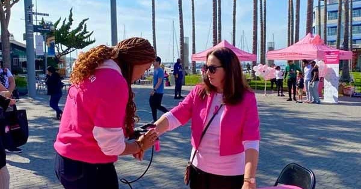 Airbrush artist in a pink shirt spraying a temporary tattoo on a guest's wrist at an outdoor waterfront event with pink tents