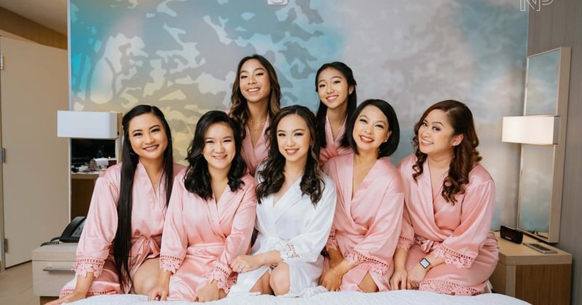 Bride in a white robe with her bridesmaids in matching pink robes on a hotel bed after on-location bridal party hair and makeup in the Bay Area
