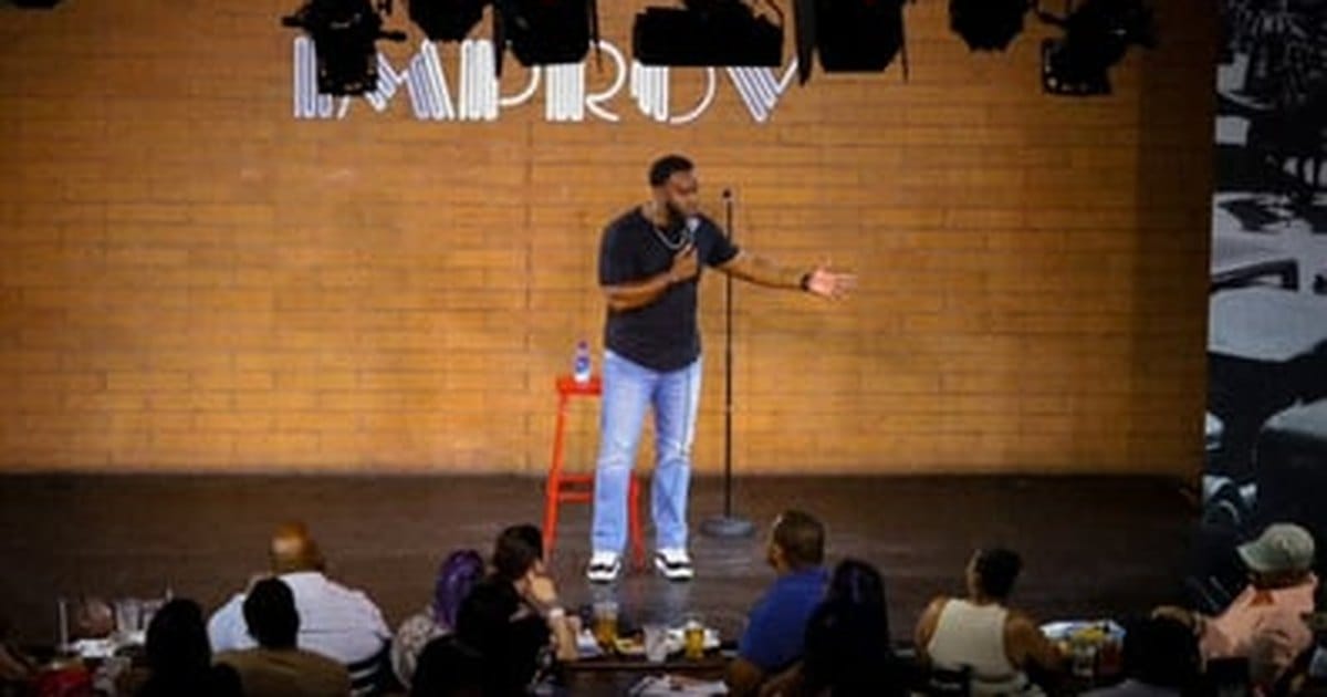 Comedian with a microphone on a brick-wall comedy stage before a seated audience, from the Elk Grove comedy show that serves the Bay Area