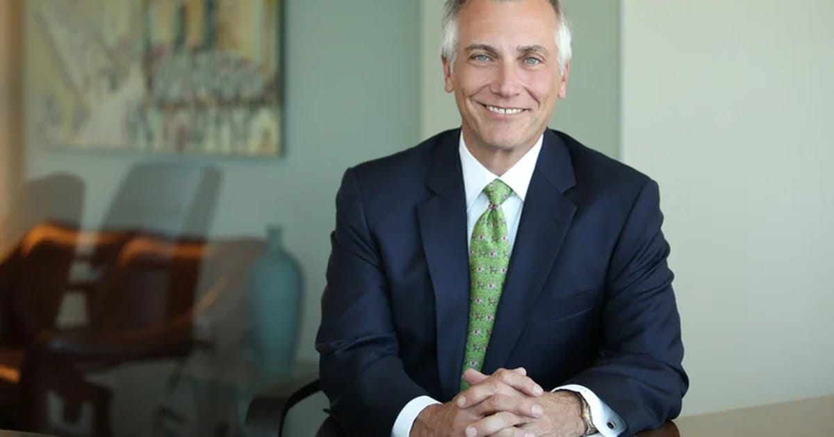 Corporate headshot of a smiling man in a navy suit and green tie at a conference table