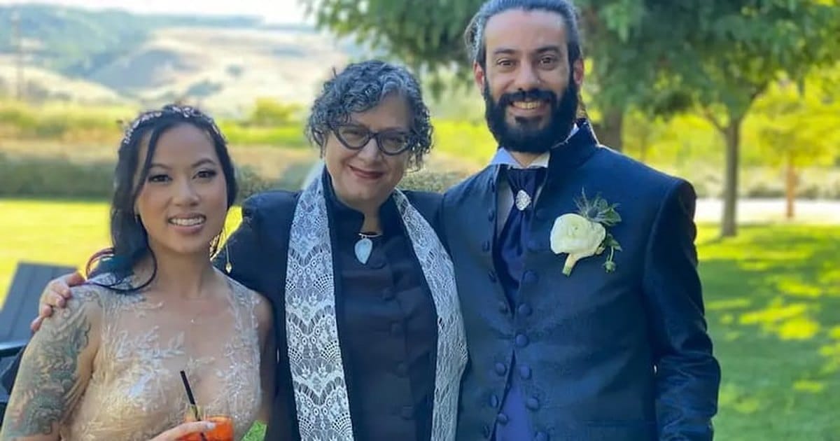 Officiant in a dark robe and white lace stole standing between a smiling bride and a groom in a navy suit on a lawn, with hills behind