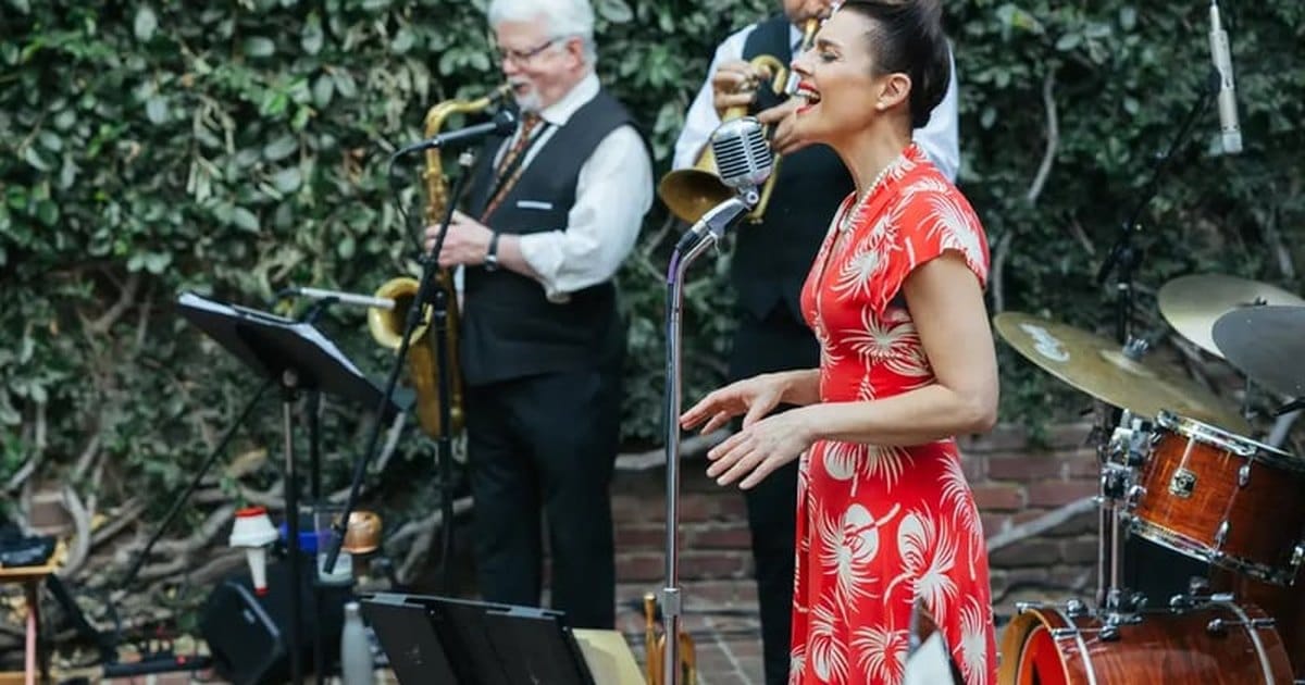 Singer in a red floral dress performing with a saxophonist and a trumpet player in front of an ivy-covered wall, from the Cosmo Alleycats Band listing, a Bay Area swing and jazz band