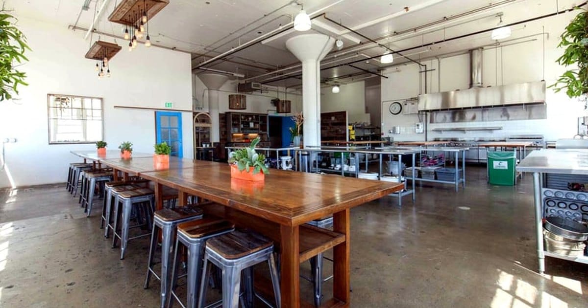 Open commercial kitchen in Dogpatch, San Francisco, with steel prep tables, a range hood and a long wooden table with metal stools