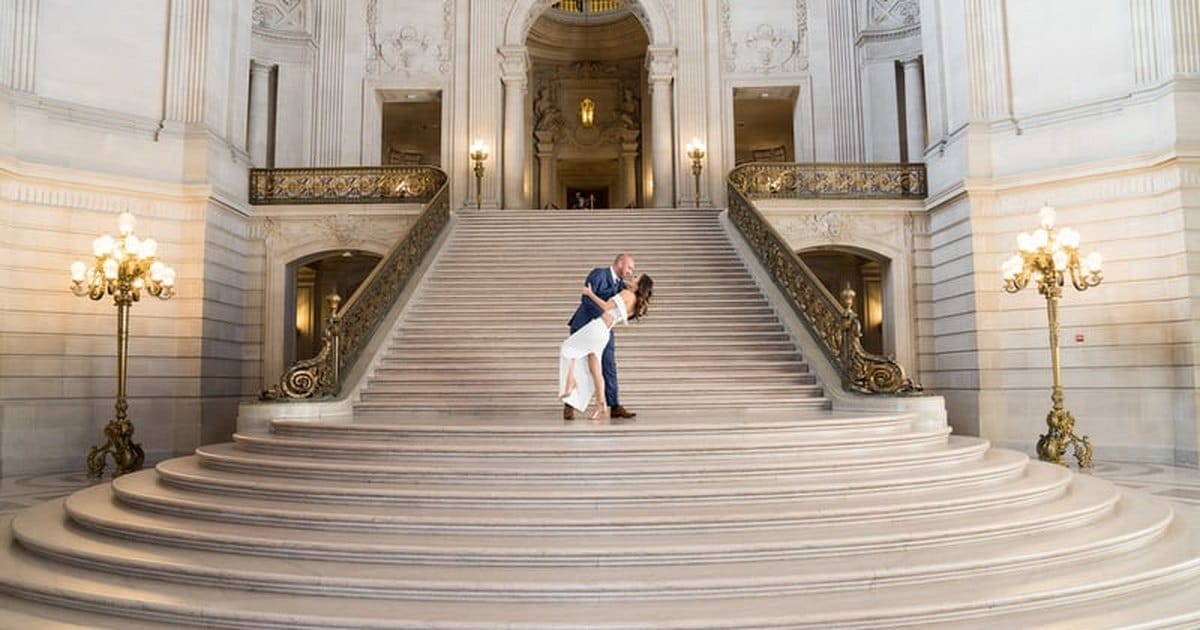 A groom dips his bride for a kiss at the top of the wide marble Grand Staircase inside San Francisco City Hall, with gold lamps on either side