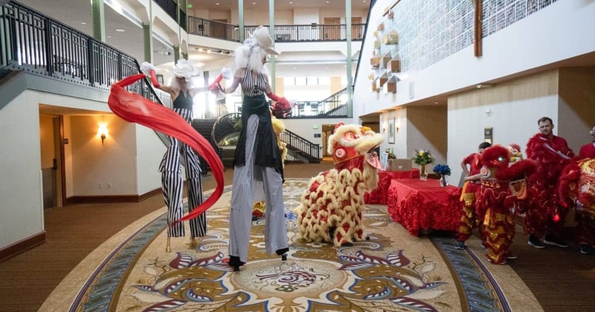 Strolling entertainers on stilts from a San Francisco listing, in striped costumes and white wigs, waving red ribbons beside a lion dance costume in an indoor atrium