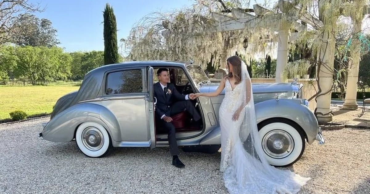 Bride and groom at the open rear door of a two-tone silver 1954 Bentley wedding car on a gravel drive
