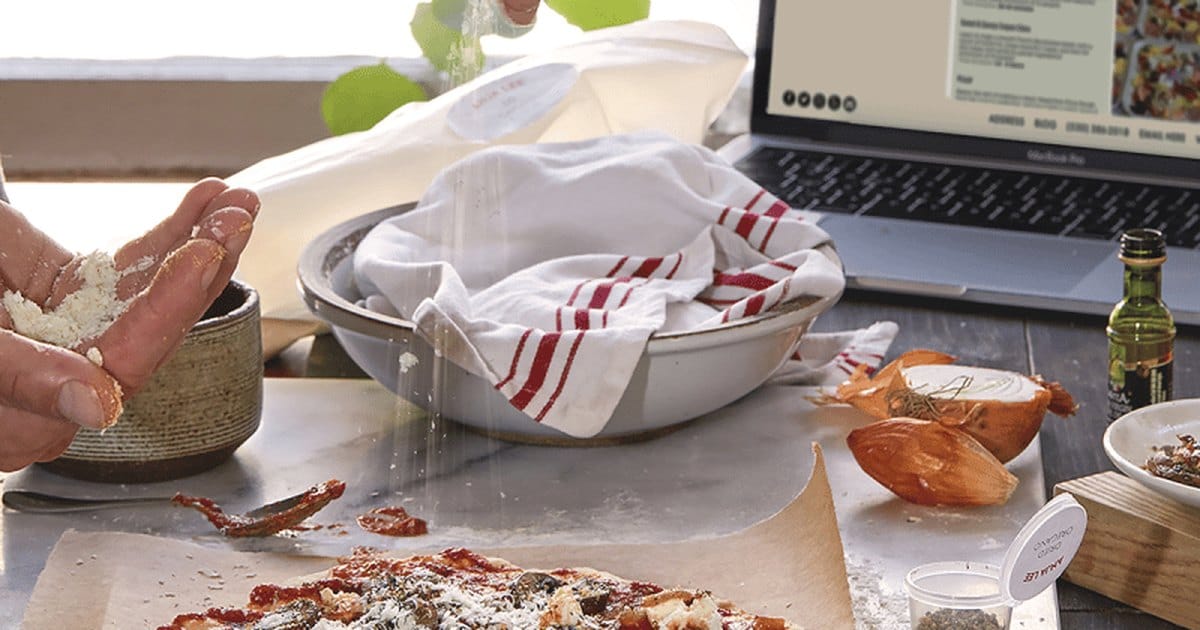 Hands sprinkling flour over a bowl of dough beside a laptop, from the Margarita Pizza with Homemade Crust virtual cooking class