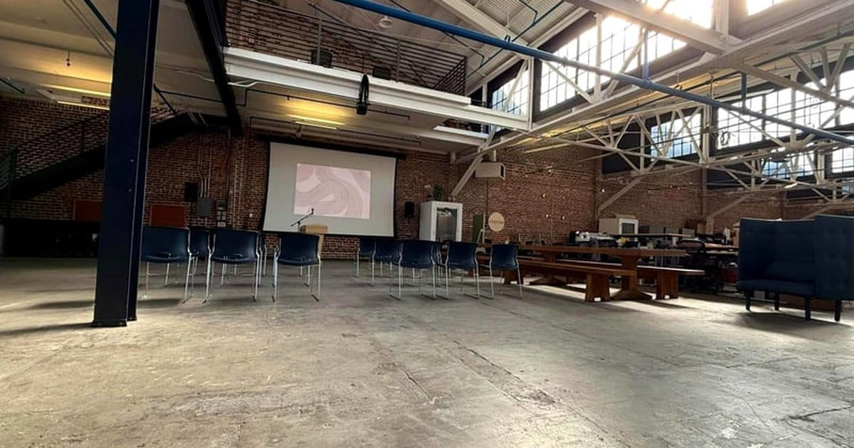 Open SoMa warehouse event space in San Francisco with a concrete floor, steel roof trusses, high windows, brick walls and a projection screen facing rows of chairs