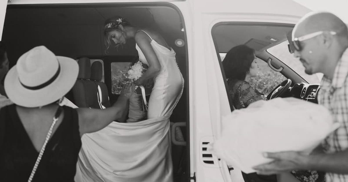 Black and white photo of a bride in a white gown stepping out of a van while guests help with her dress, from a San Francisco wedding content creator package