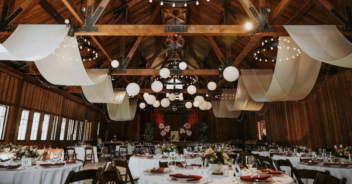 Barn wedding reception set with round tables, white draped beams, paper lanterns and string lights, from the Elopement and Microwedding Planning and Coordination listing