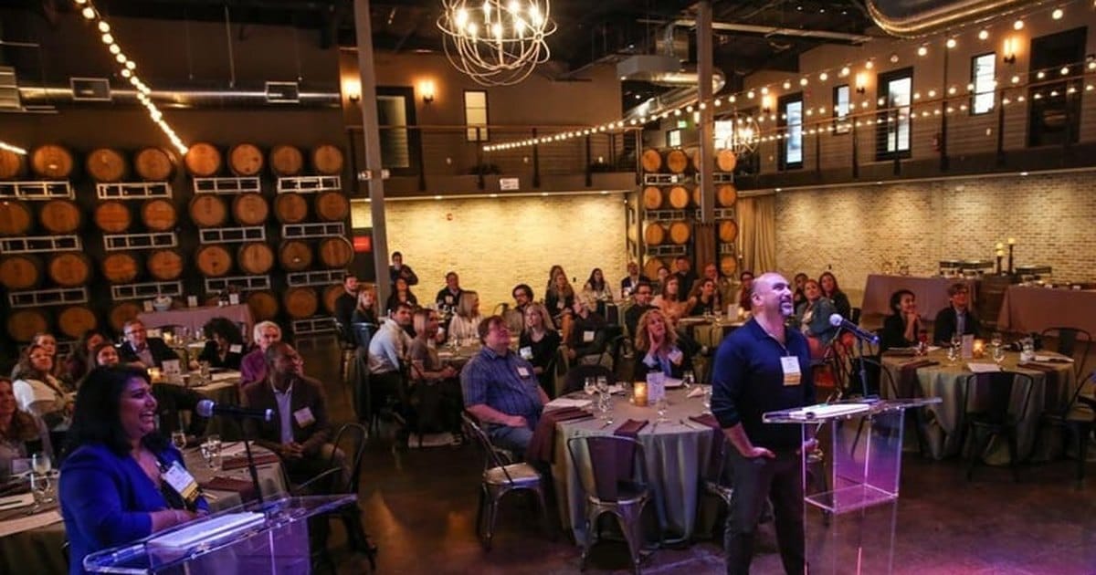 Two speakers at clear podiums addressing guests at round tables in a San Francisco winery event space with barrel racks and string lights