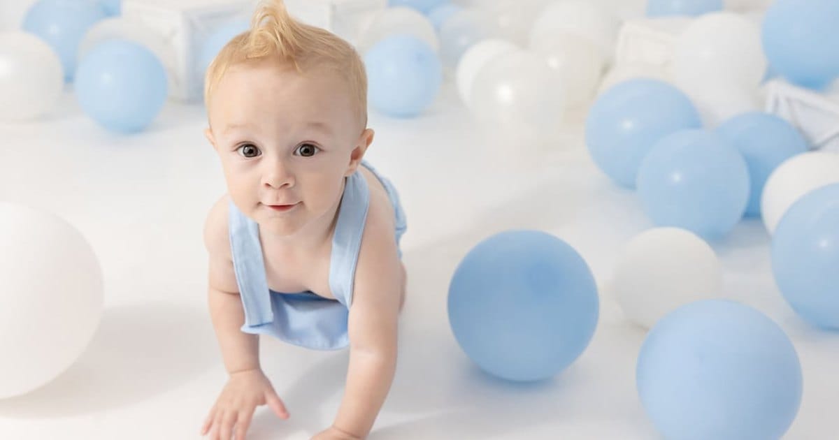 Baby in light blue overalls crawling across a white studio floor among scattered blue and white balloons