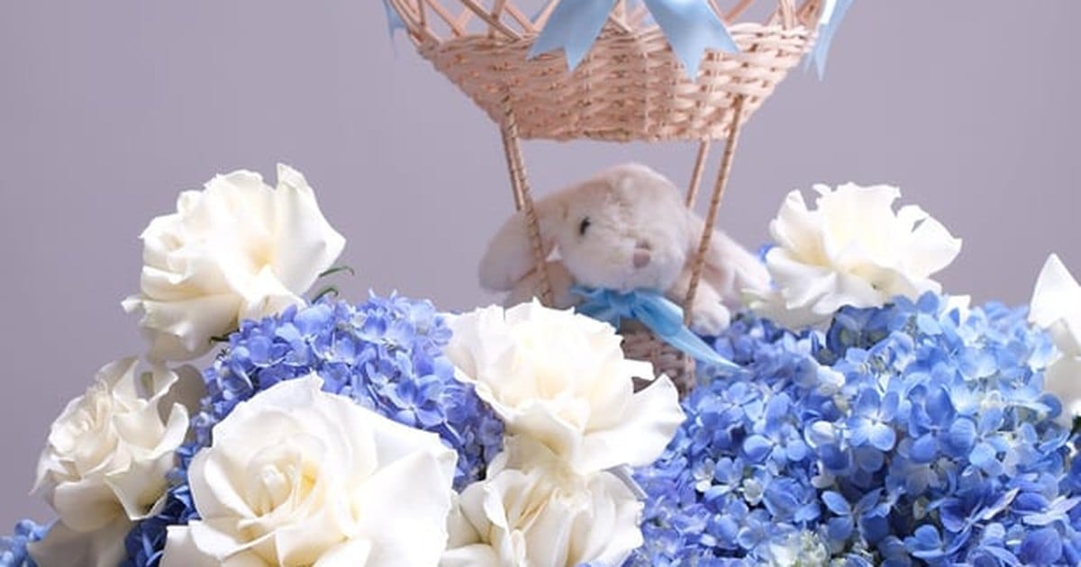 Blue hydrangeas and white roses around a plush rabbit in a woven hot air balloon, a baby shower arrangement from a San Francisco florist