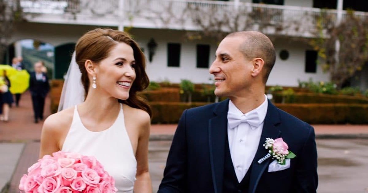 Smiling bride with auburn hair, drop earrings, a veil and pink roses beside a groom in a navy suit, white bow tie and rose boutonniere, in front of a white building