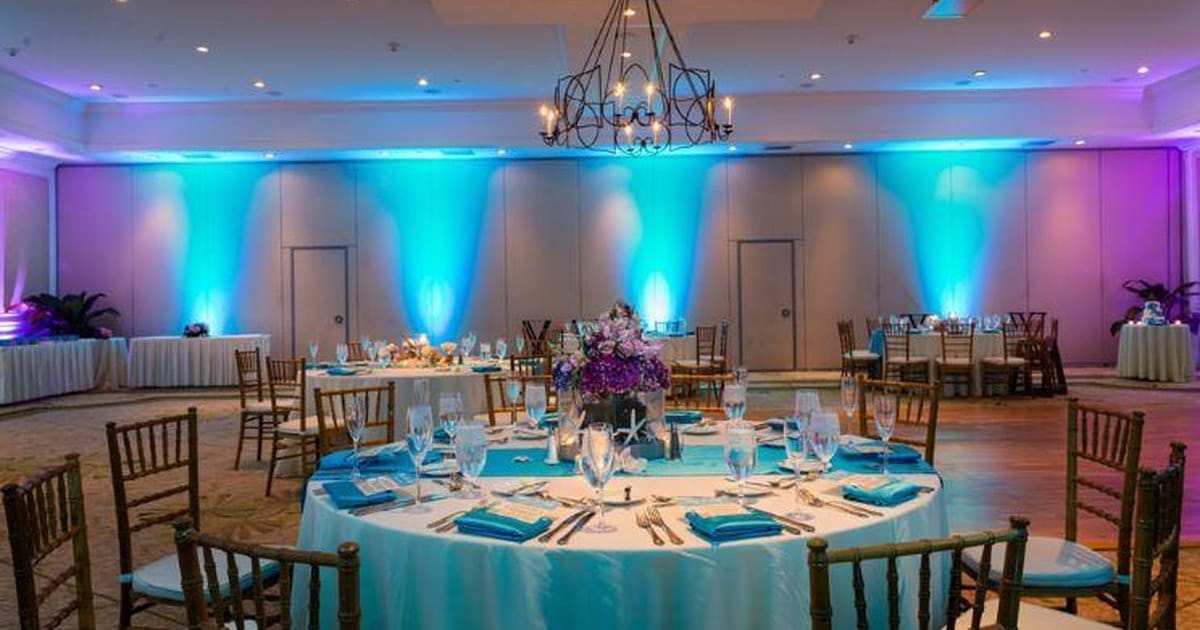 Hotel ballroom with teal and violet uplighting on the walls, an iron chandelier and round tables set with turquoise napkins and purple flowers