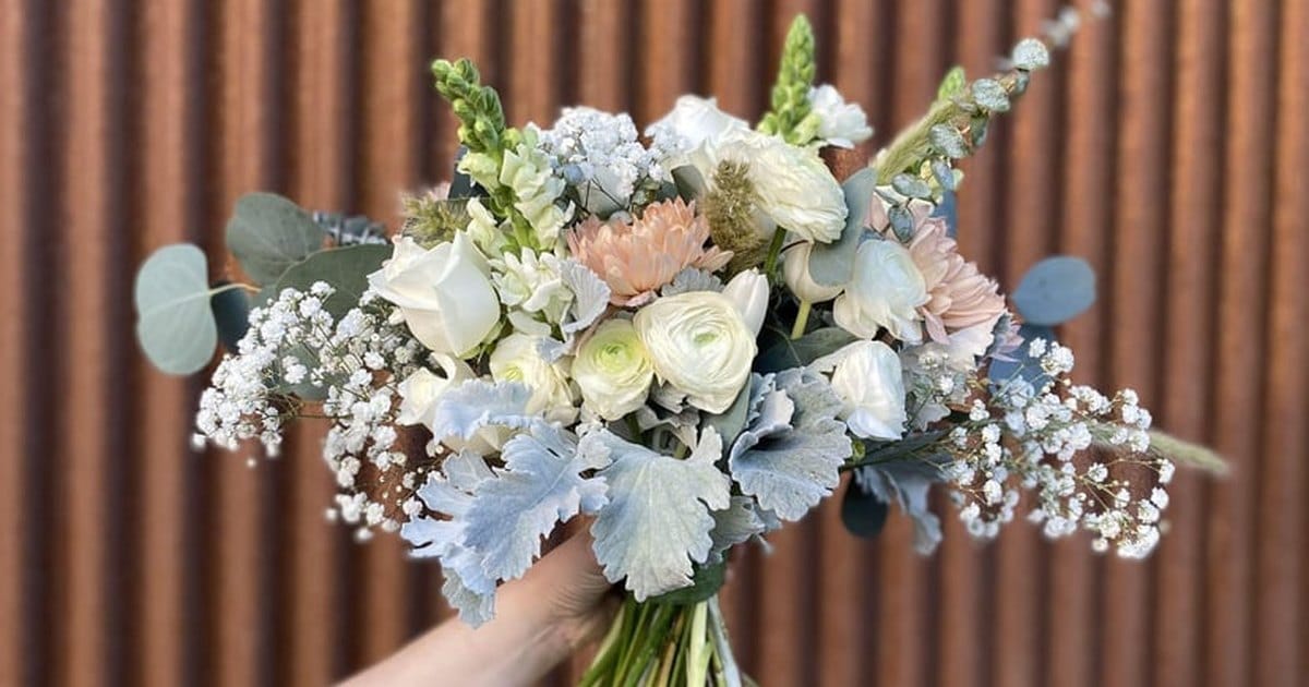 Hand holding up a loose bridal bouquet of white ranunculus and roses, peach dahlias, baby's breath and silver dusty miller against a rusted metal wall