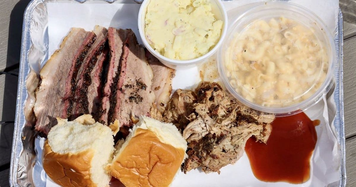 Foil tray of sliced brisket, pulled pork, potato salad, mac and cheese, two soft rolls and barbecue sauce on a wooden deck