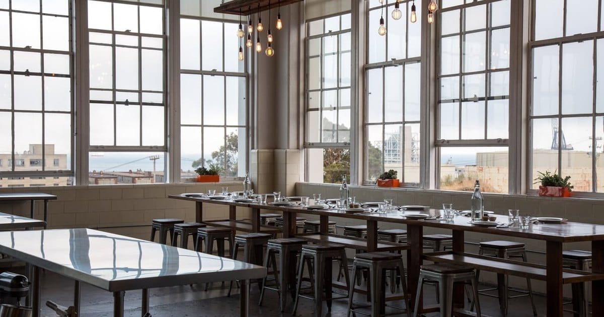 Empty dining area with a long wooden table set with plates and glasses, metal stools, hanging bulb lights and tall factory windows