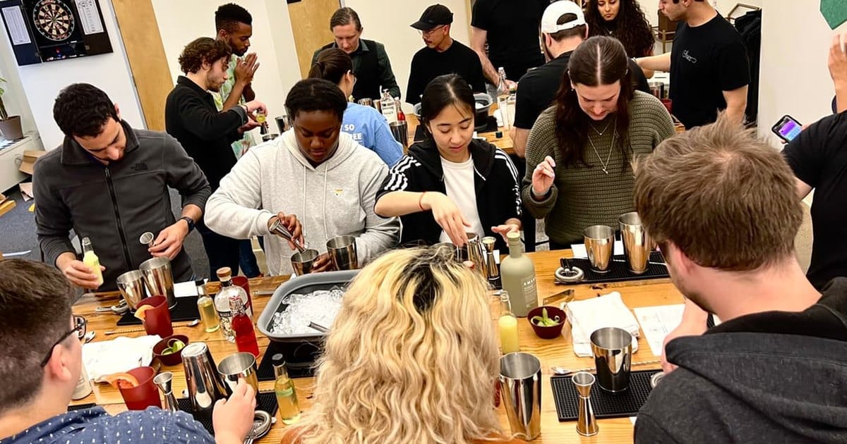 Coworkers crowded around long wooden tables measuring and shaking cocktails with steel shakers, jiggers and a tub of ice