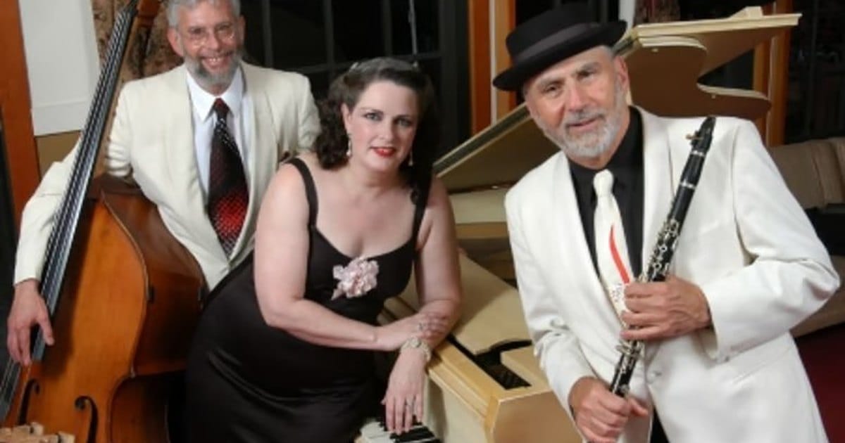 Upright bass player, a singer in a black dress at a white grand piano and a clarinetist in a white jacket and black hat posing together