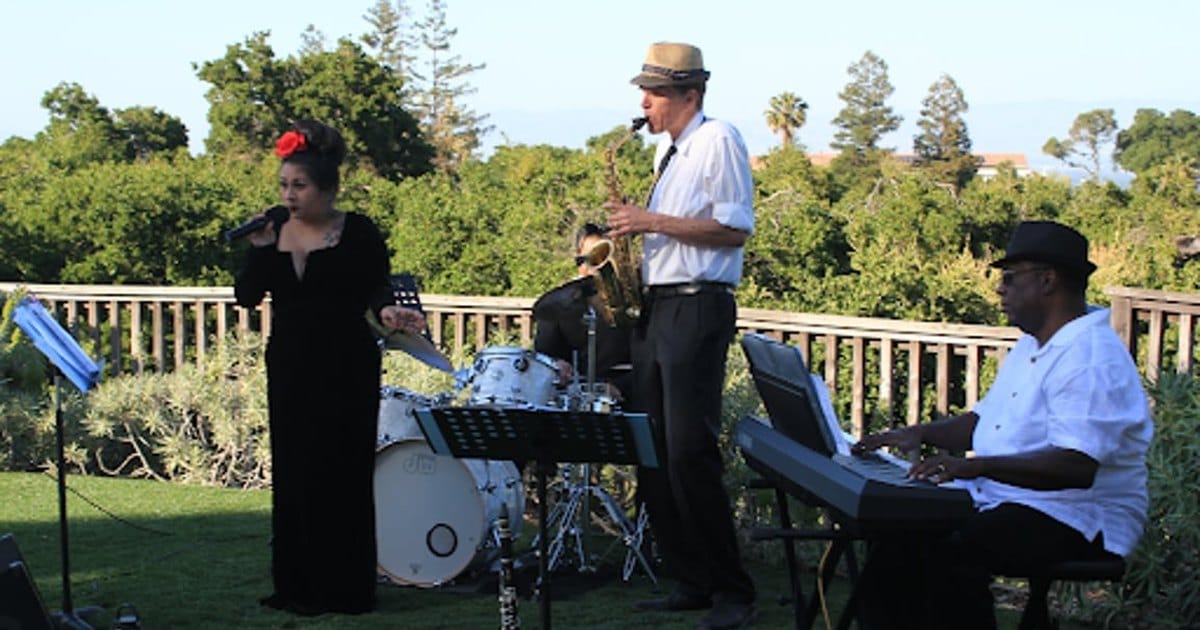 Singer in a black gown, a saxophonist in a straw hat, a drummer and a keyboard player performing outdoors on a lawn beside a wooden railing