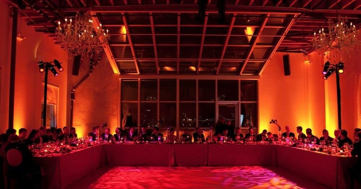 Long U-shaped dinner table under a glass skylight roof, with crystal chandeliers, amber uplighting and a patterned light projection on the floor