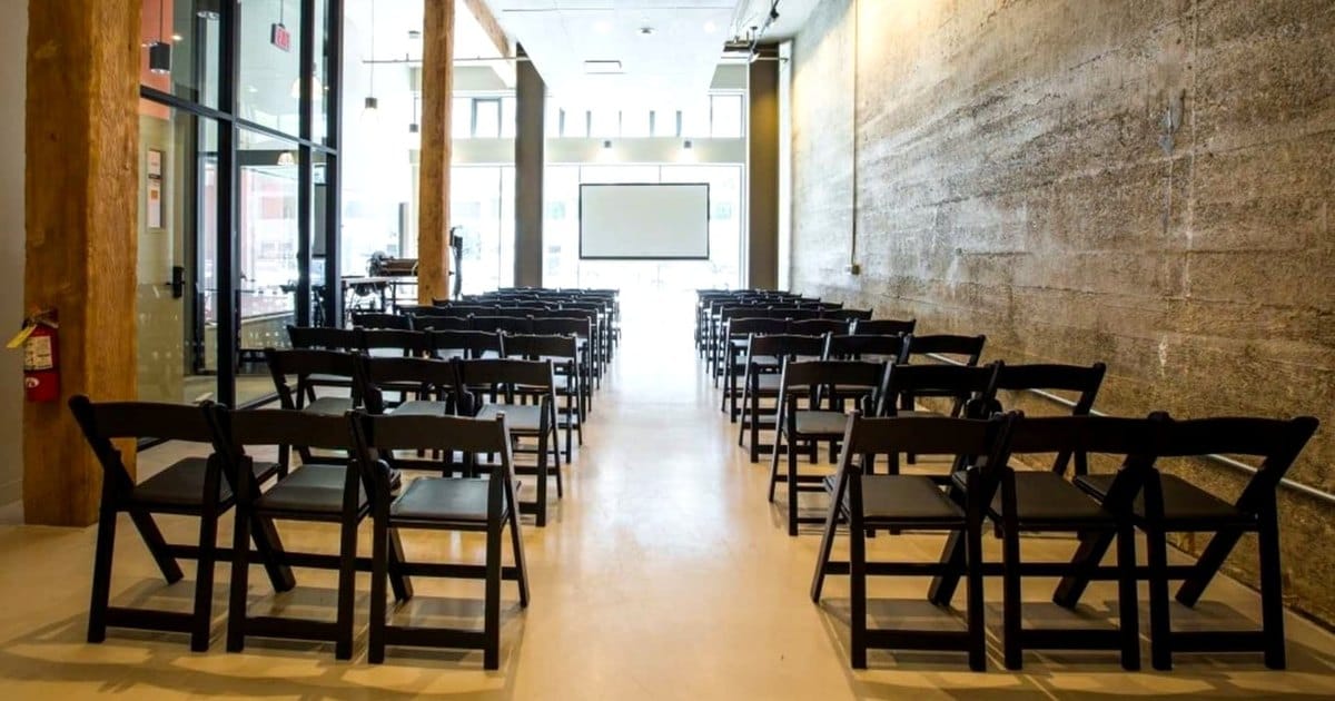 Rows of black folding chairs facing a projection screen in a long event room with a concrete wall, timber posts and a glass storefront