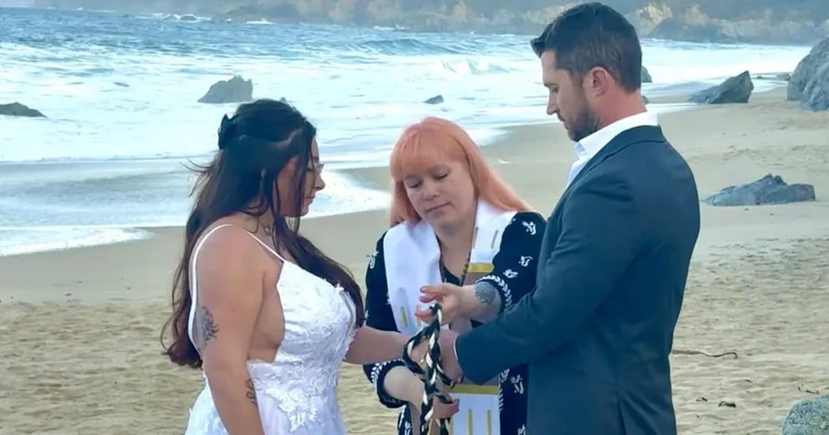 Officiant wrapping a cord around a couple's joined hands in a handfasting on a beach, with waves and rocks behind them