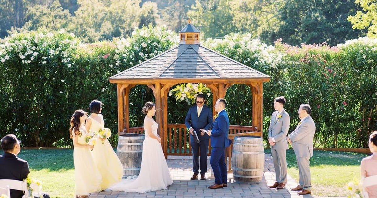 Outdoor wedding ceremony at a wooden gazebo with a bride in white, bridesmaids in yellow and groomsmen in gray, wine barrels beside the steps