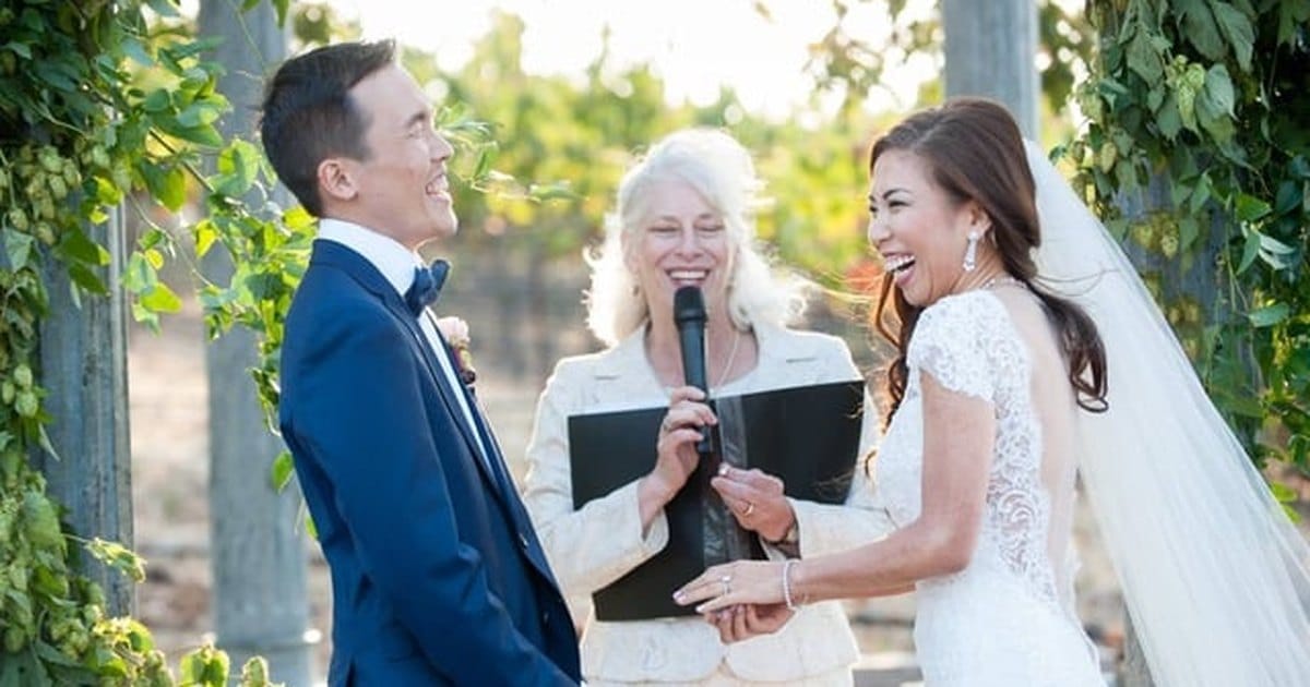 Laughing bride and groom holding hands in front of a smiling officiant with a microphone and black folder, framed by leafy vines outdoors
