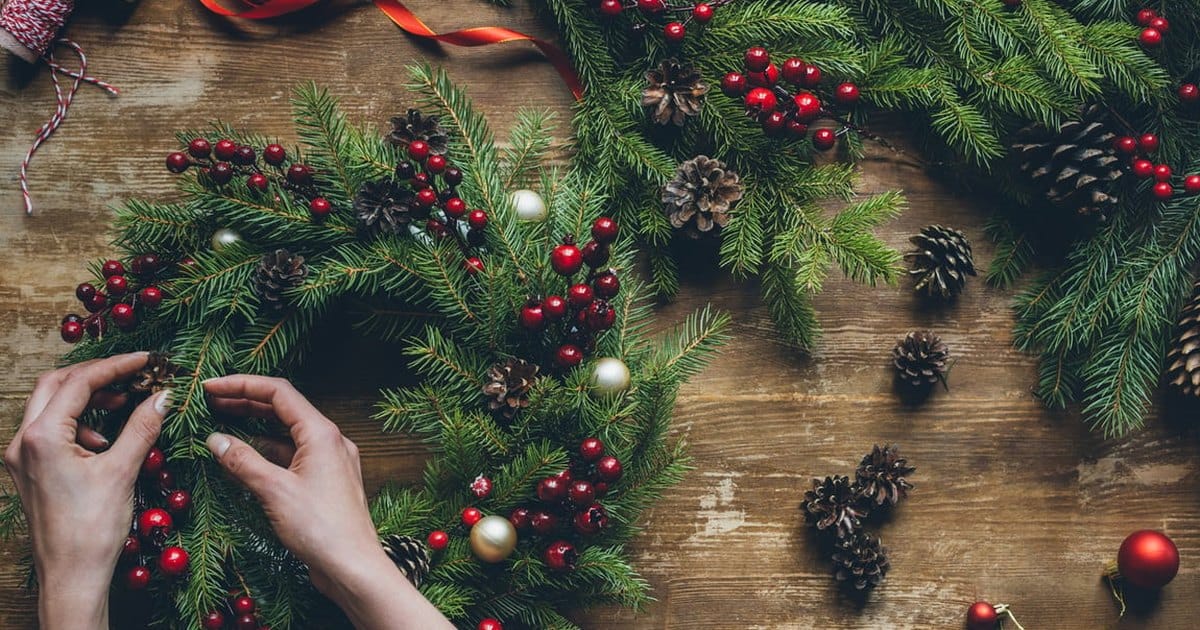 Hands arranging a fresh fir wreath with red berries, pine cones and small ornaments on a wooden table, with red ribbon and twine nearby