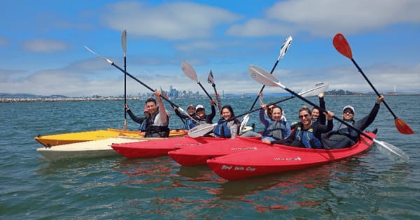 A team in red and yellow kayaks raising their paddles on a guided Alameda kayak tour, with the San Francisco skyline in the distance