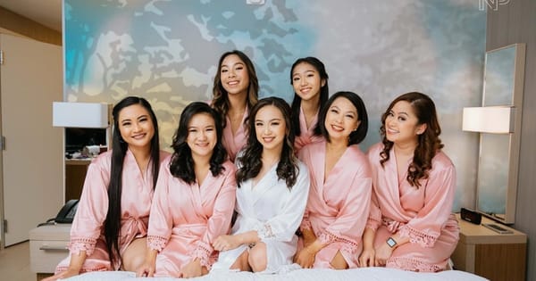 Bride in a white robe with her bridesmaids in matching pink robes on a hotel bed after on-location bridal party hair and makeup in the Bay Area