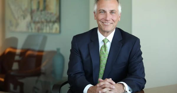 Corporate headshot of a smiling man in a navy suit and green tie at a conference table