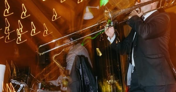 A trombone player in a dark suit performs in front of a saxophonist under warm string lights at an evening reception, from an Oakland DJ and live sax hybrid act