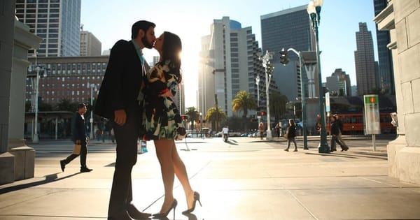 Engagement photo of a couple kissing on a sunny downtown San Francisco sidewalk with high-rises, palm trees and a streetcar behind them
