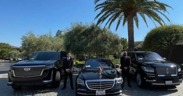 Two chauffeurs in dark suits beside a black sedan and two black SUVs parked under a palm tree