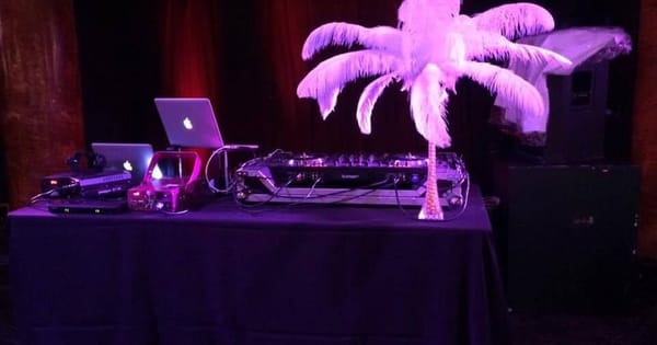 Foster City event DJ table with two laptops, a mixing controller and a white feather centerpiece under purple stage lighting, with speakers stacked behind