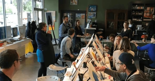 An instructor talks to a group seated at easels along a long table during a paint and sip team building class