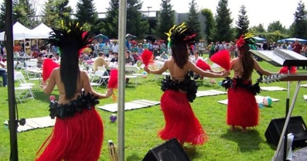 Hula dancers for a luau party in red grass skirts and feathered headdresses performing on a lawn in front of seated guests at an outdoor event