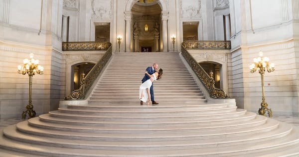 A groom dips his bride for a kiss at the top of the wide marble Grand Staircase inside San Francisco City Hall, with gold lamps on either side