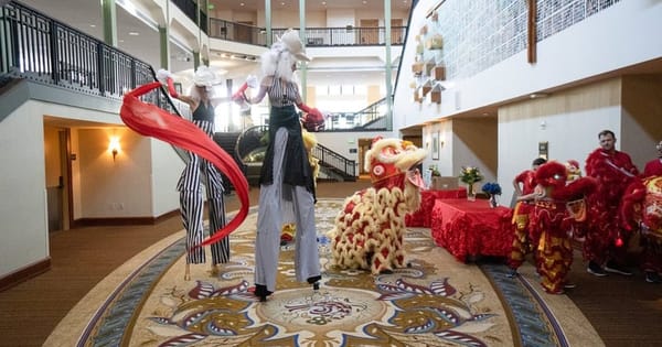 Strolling entertainers on stilts from a San Francisco listing, in striped costumes and white wigs, waving red ribbons beside a lion dance costume in an indoor atrium
