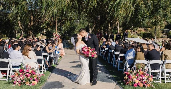 Newlyweds kissing on a stone garden aisle lined with pink flower arrangements and seated guests under willow trees