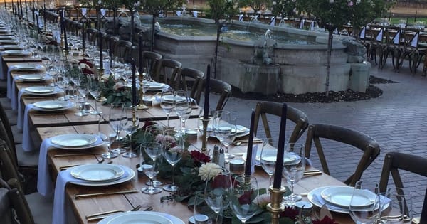 Long outdoor dinner table set for plated service with white plates, candles and a floral runner beside a vineyard fountain, from a Sonoma caterer