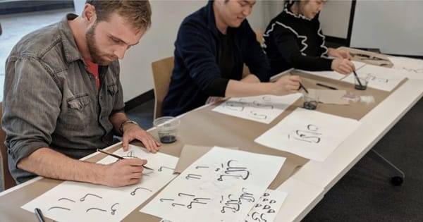 Three colleagues practicing Persian and Arabic calligraphy with pens and ink at a long table in an office workshop