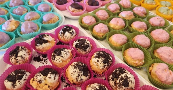 Trays of mini cream puffs in pink, green, teal and yellow paper liners, topped with cookie crumbs, pink, purple and green glazes and chocolate