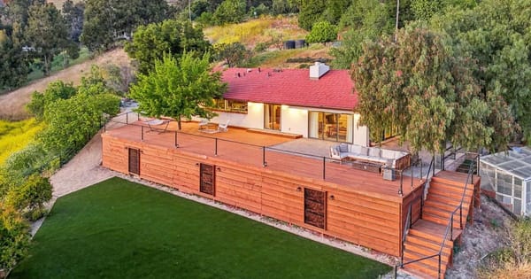 Aerial view of a red-roofed hillside home with a long wooden deck, patio seating and a green lawn below, framed by trees and grassy hills