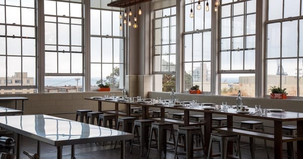 Empty dining area with a long wooden table set with plates and glasses, metal stools, hanging bulb lights and tall factory windows