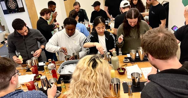 Coworkers crowded around long wooden tables measuring and shaking cocktails with steel shakers, jiggers and a tub of ice