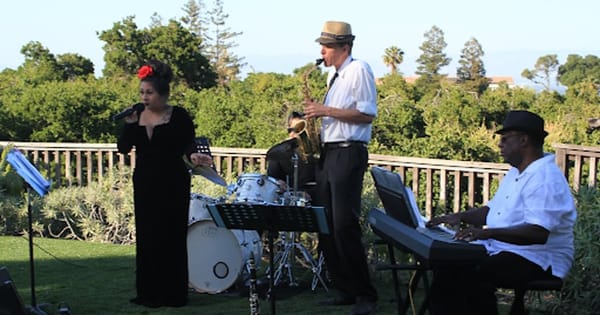 Singer in a black gown, a saxophonist in a straw hat, a drummer and a keyboard player performing outdoors on a lawn beside a wooden railing